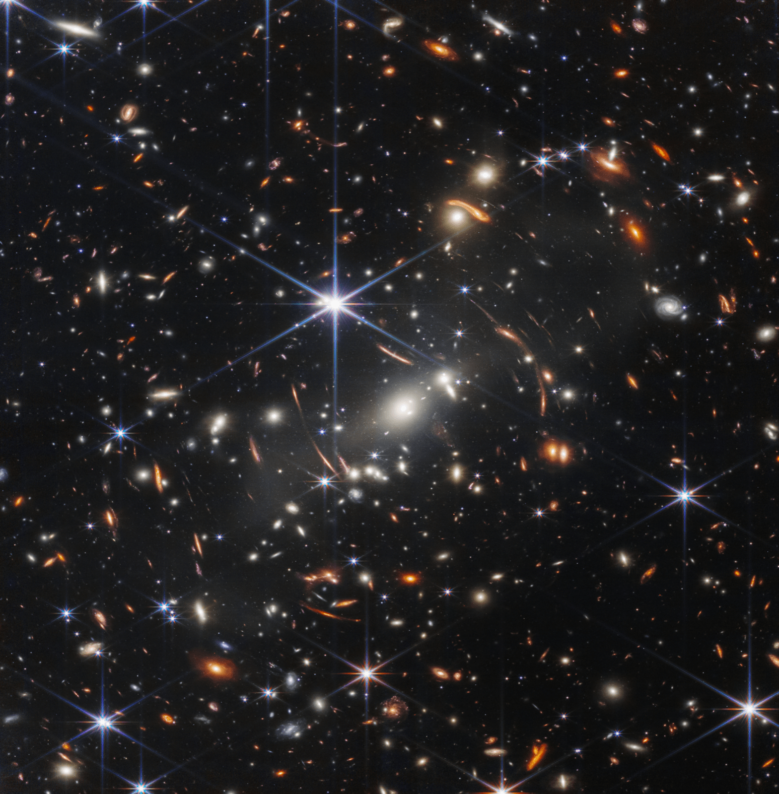 In this image, captured by JWST, has a background of space is black. Thousands of galaxies appear all across the view. Their shapes and colors vary. Some are various shades of orange, others are white. Most stars appear blue, and are sometimes as large as more distant galaxies that appear next to them. A very bright star is just above and left of center. It has eight bright blue, long diffraction spikes. Between 4 o’clock and 6 o’clock in its spikes are several very bright galaxies. A group of three are in the middle, and two are closer to 4 o’clock. These galaxies are part of the galaxy cluster SMACS 0723, and they are warping the appearances of galaxies seen around them. Long orange arcs appear at left and right toward the center.