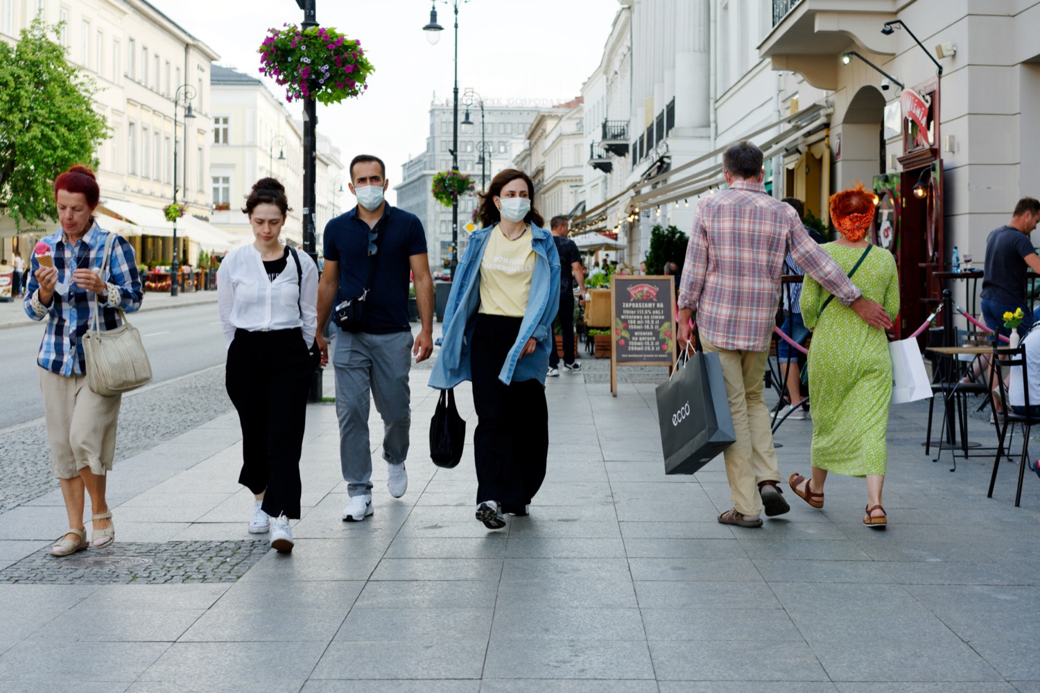 six white people walking on a european street. four people are walking towards the camera, two of which are wearing masks, and the two people not facing the camera walking away, appearing to not be wearing masks