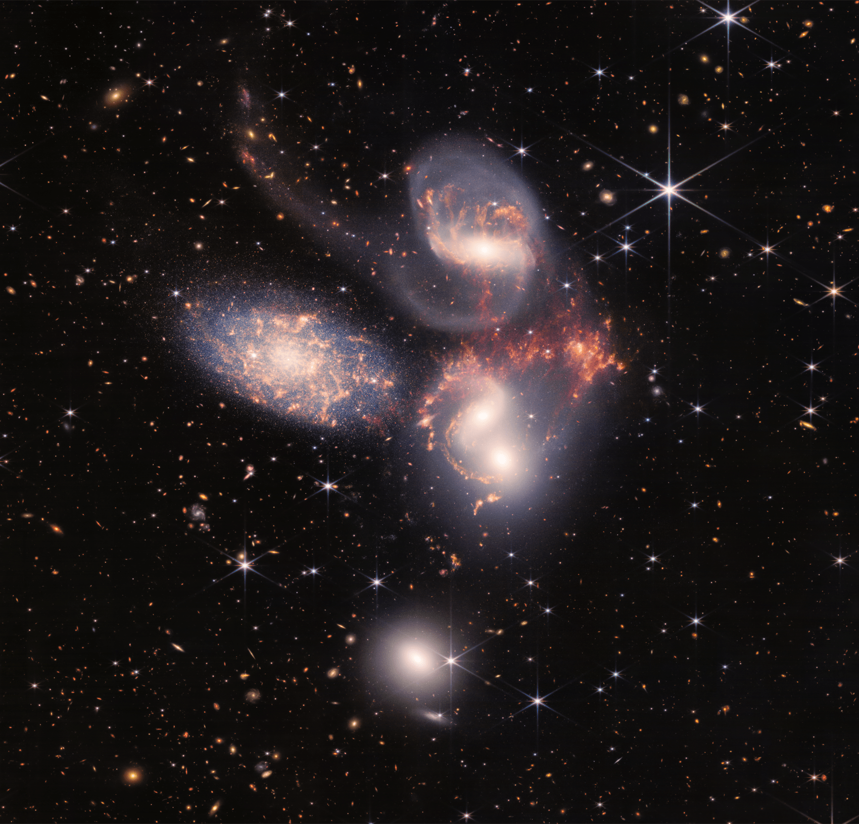 An image, captured by JWST, of a group of five galaxies that appear close to each other in the sky: two in the middle, one toward the top, one to the upper left, and one toward the bottom. Four of the five appear to be touching. One is somewhat separated. In the image, the galaxies are large relative to the hundreds of much smaller (more distant) galaxies in the background. All five galaxies have bright white cores. Each has a slightly different size, shape, structure, and coloring. Scattered across the image, in front of the galaxies are number of foreground stars with diffraction spikes: bright white points, each with eight bright lines radiating out from the center.