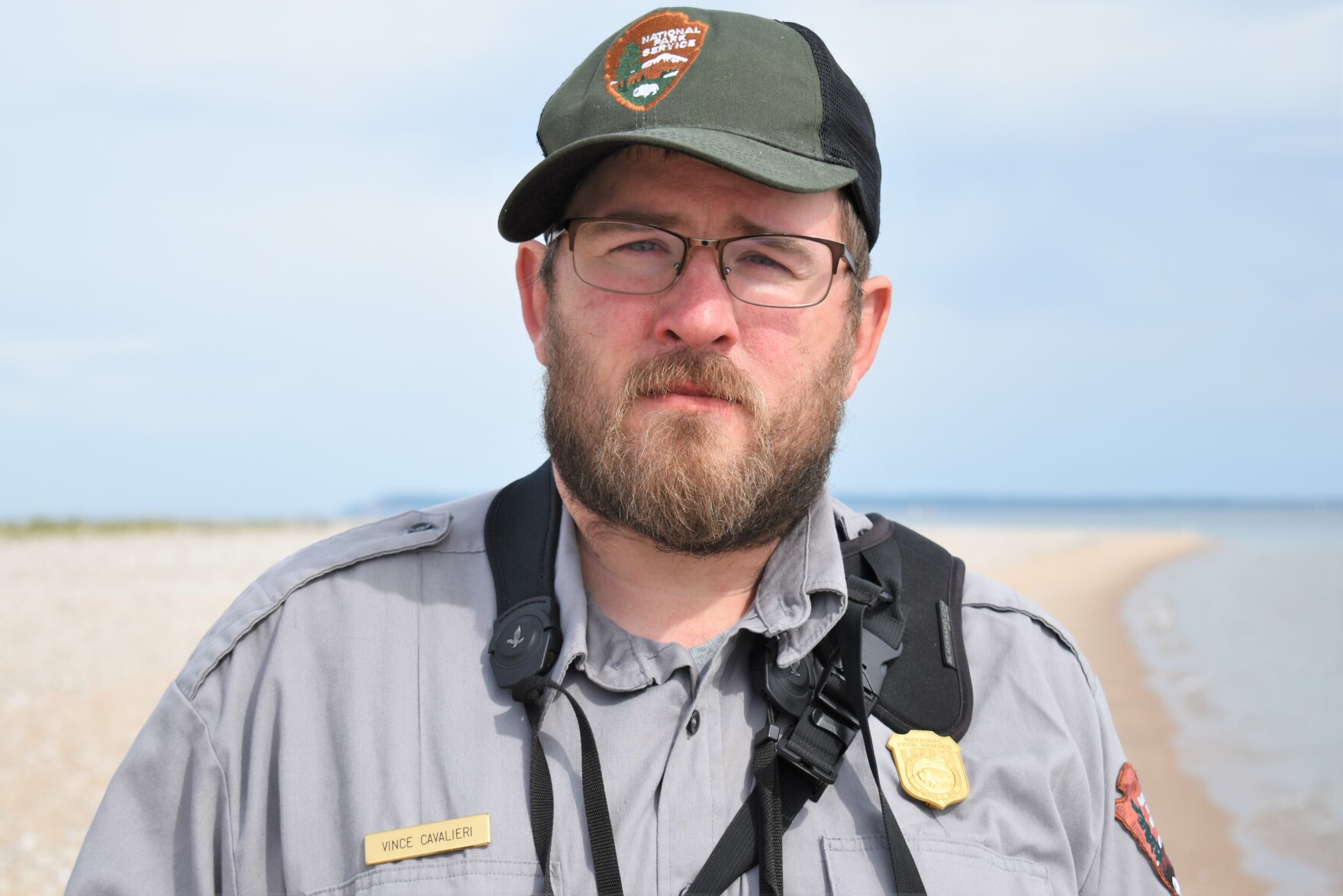a white man with a beard and glasses wearing a park uniform looks solemnly into the camera