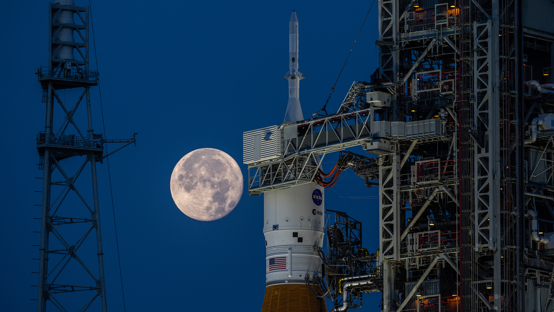 zoomed-in photograph of an unmanned rocket on the launch pad showing just the top portion, with the moon looming in the background