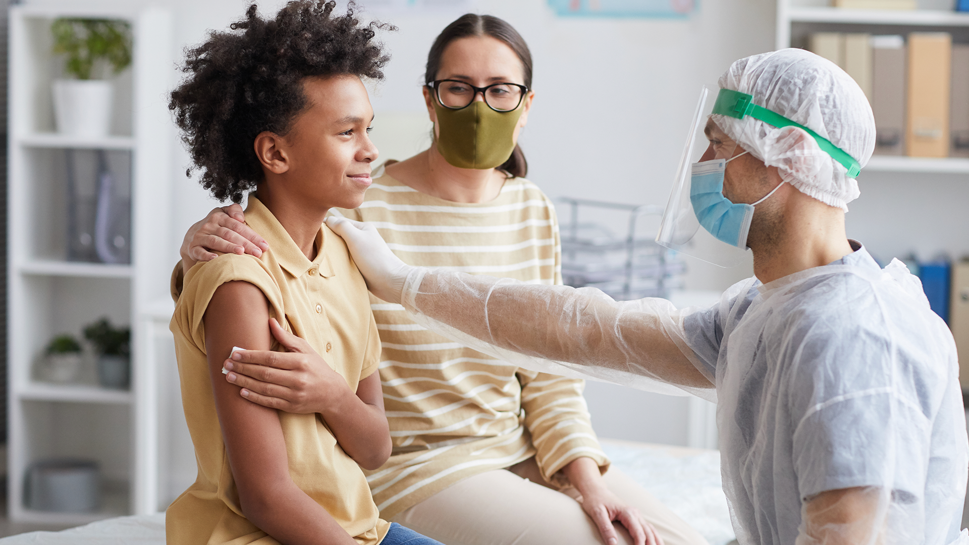 photo of a woman and middle school-aged child sitting side by side at a doctor's office, with a doctor reaching over to kindly touch the child's arm, while the child holds a piece of gauze to this other arm and smiles slightly
