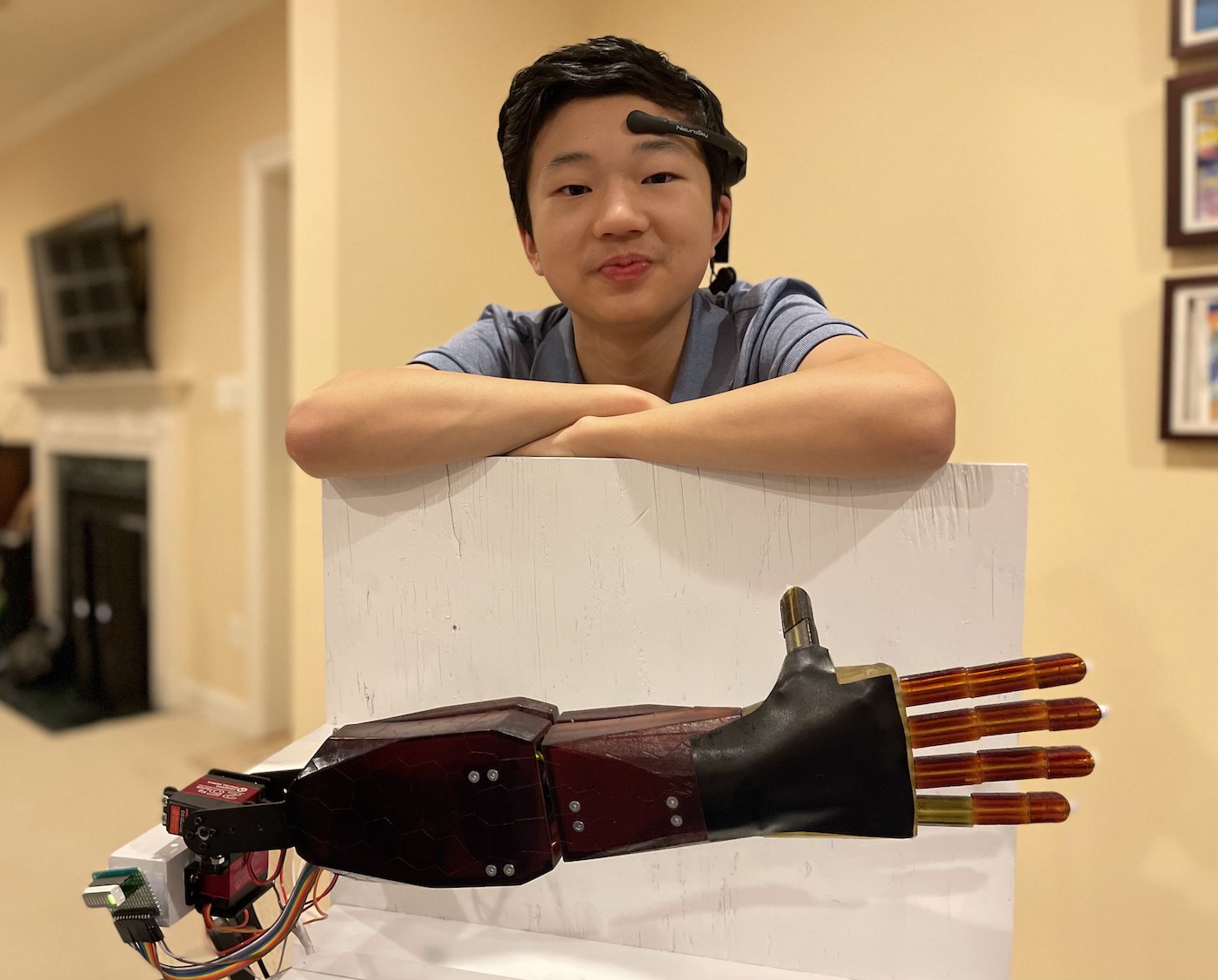 A teenage boy looks at the camera with a clever grin, wearing a device that looks like a telecommunication headset pressed to his forehead. He rests his elbows on a white stand, in front of which lies a robotic arm.