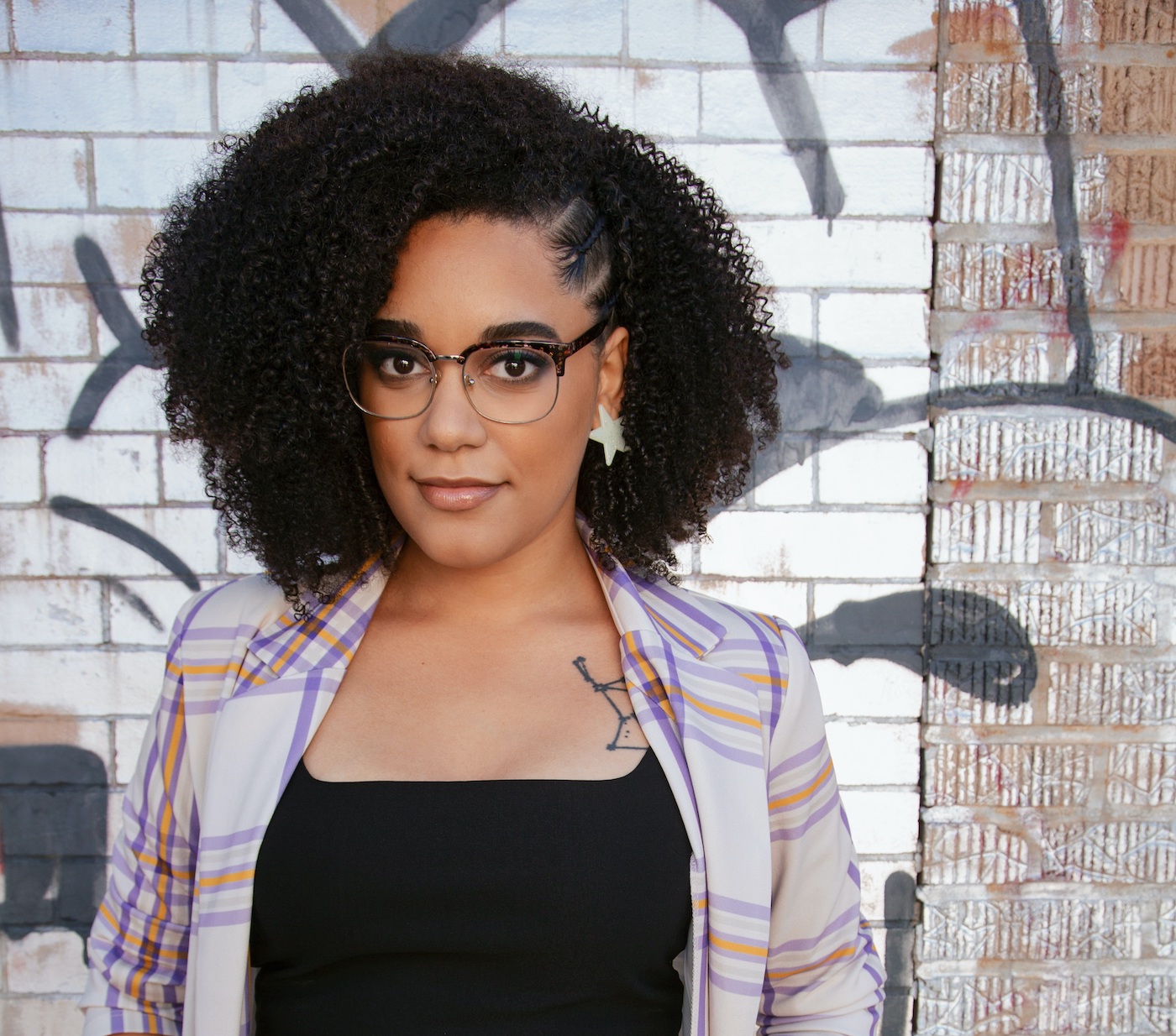 A woman with glasses looks into the camera with a thoughtful, gentle smile. A tattoo of a constellation peeks out from the right side of her shirt, by her collarbone.