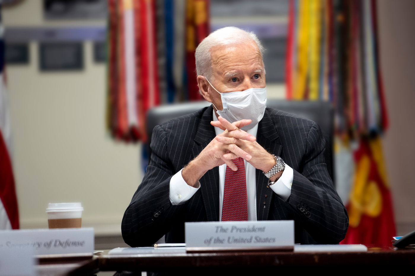 Joe Biden with a face mask on sitting at a desk with his hands folded in front of him. A placard in the foreground reads "The President of the United States"