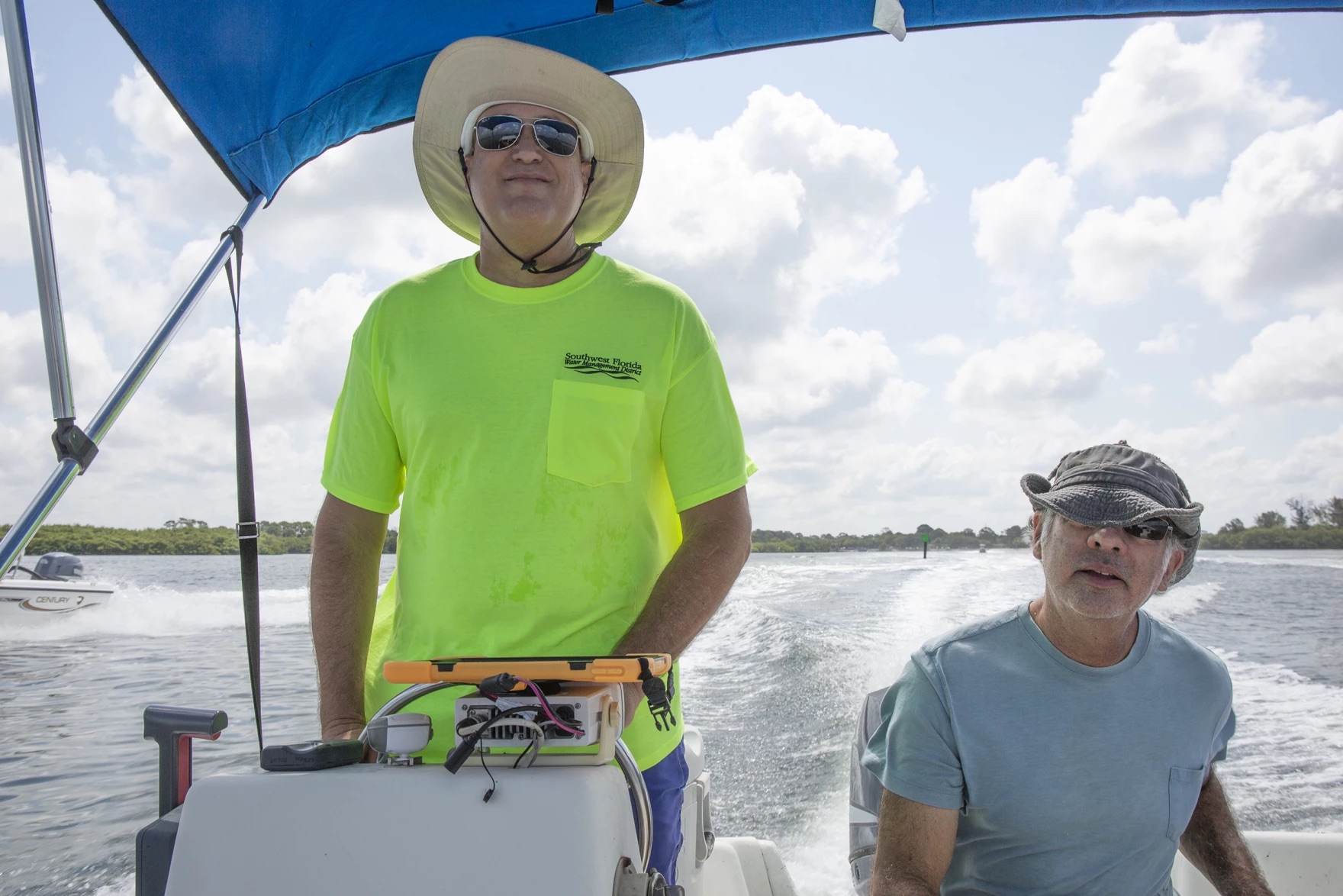 two older white men on a boat, both wearing hats and sunglasses. the one on the left is steering the boat, and there's a wake of the boat's water in the background