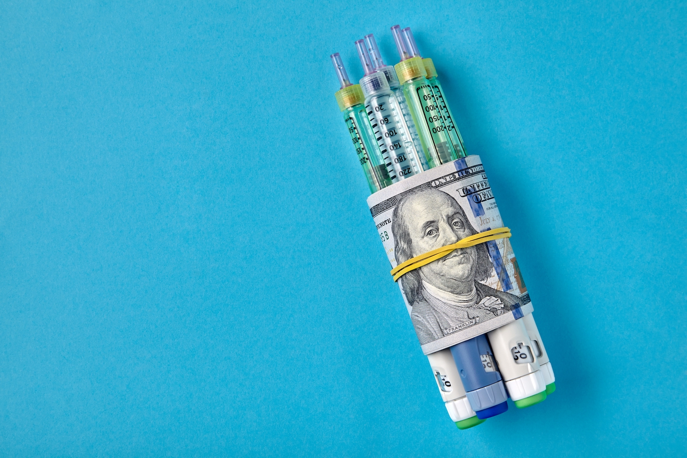 Insulin syringes wrapped in hundred dollar bills, blue background.