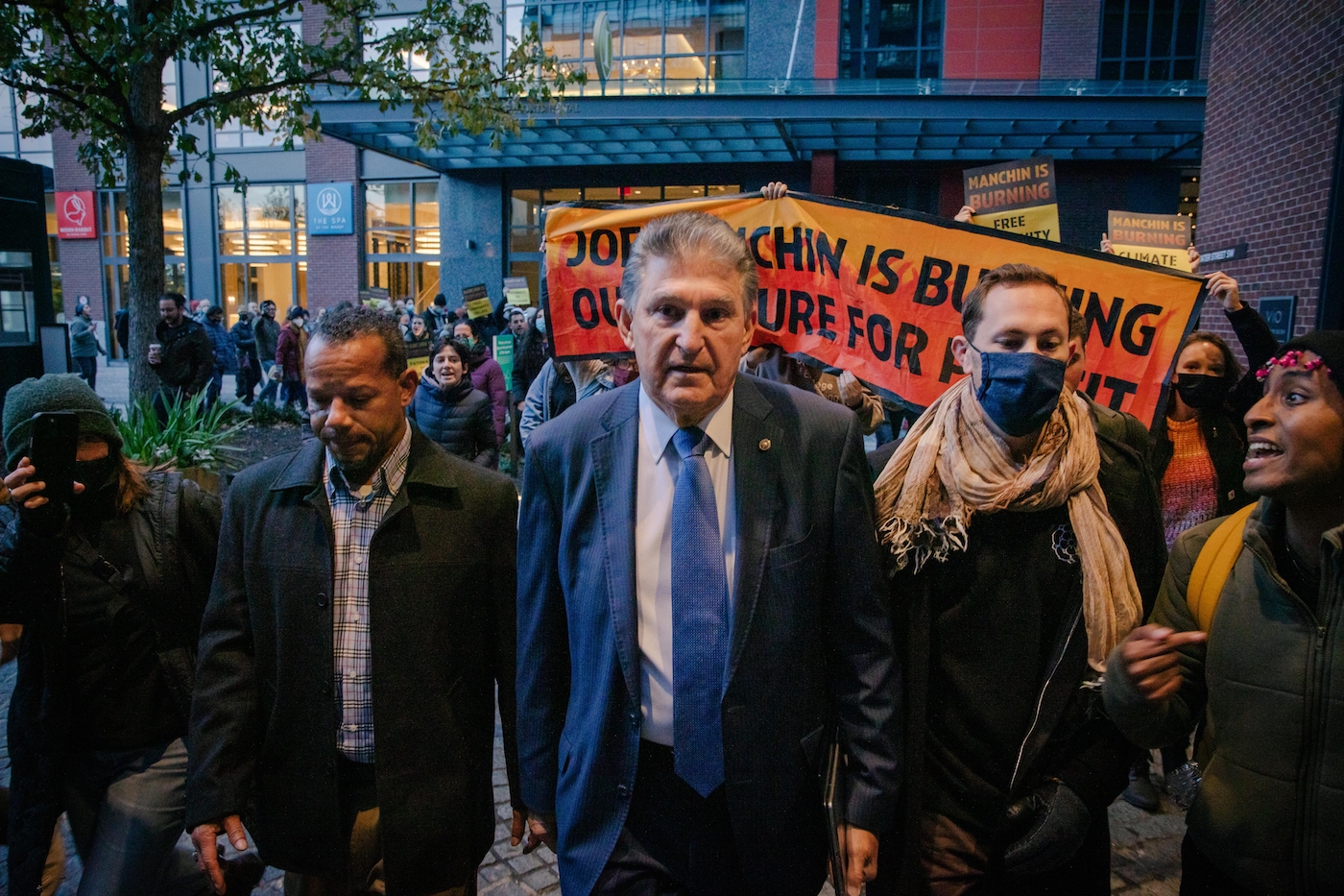 Senator Joe Manchin walking towards the camera as protesters to his left and right make their voices heard. A large sign behind Manchin is partially obscured, but reads "Joe Manchin is Burning Our Future For Profit."