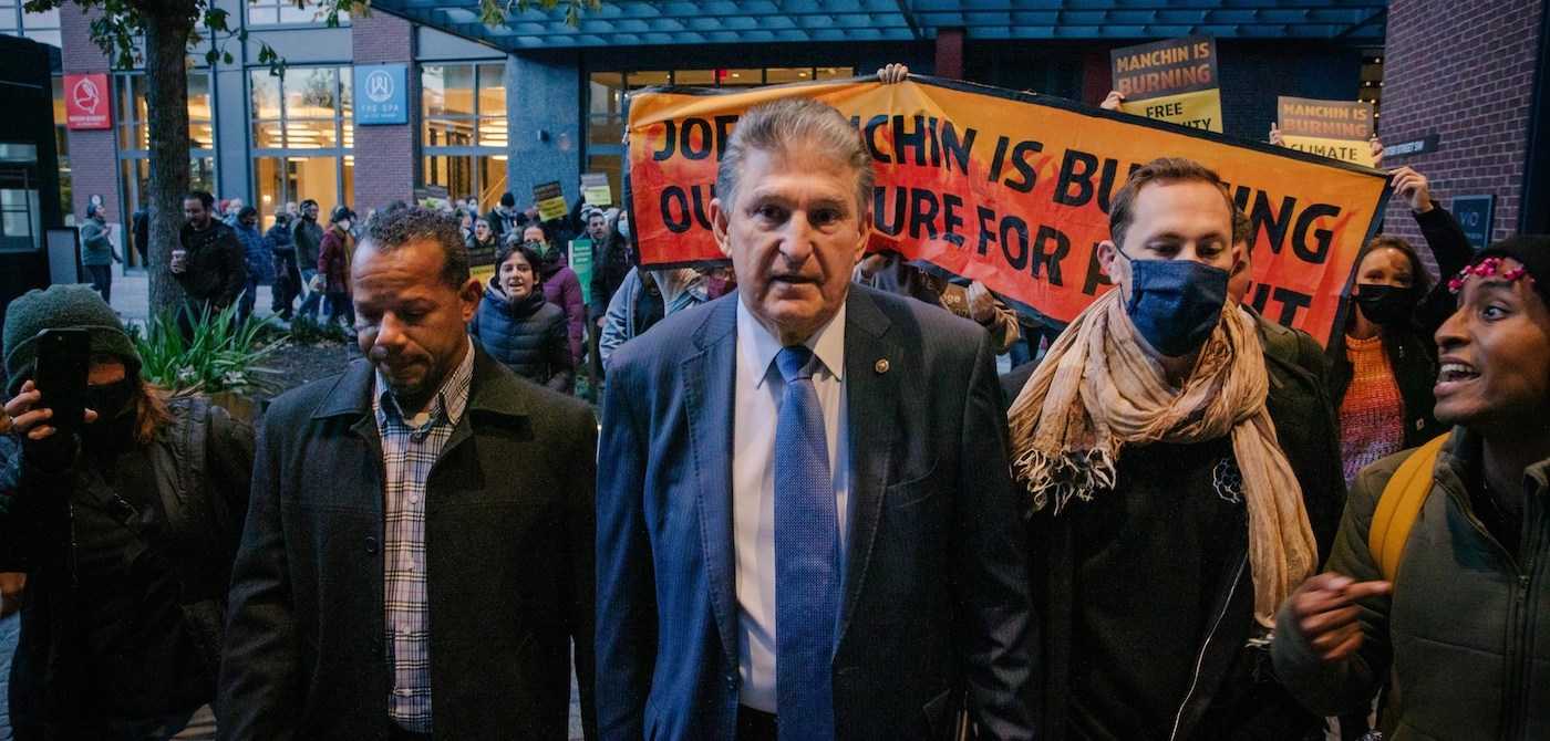 Senator Joe Manchin walking towards the camera as protesters to his left and right make their voices heard. A large sign behind Manchin is partially obscured, but reads "Joe Manchin is Burning Our Future For Profit."