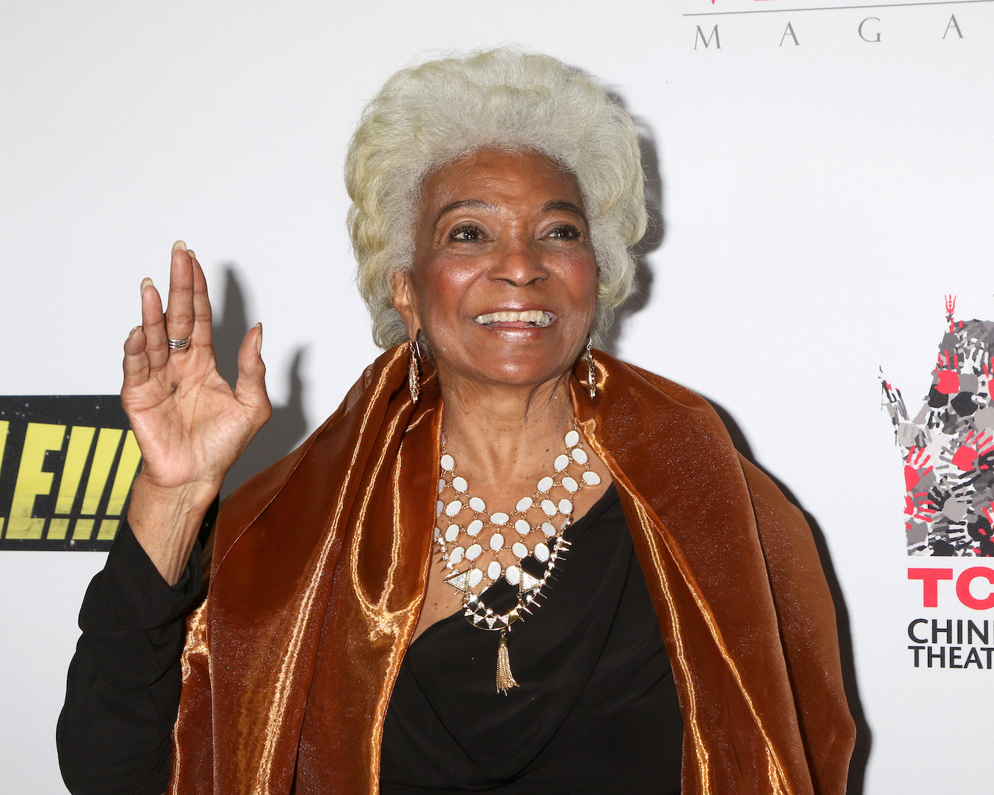 Nichelle Nichols at a premiere, wearing a black shirt, a caramel colored velvet shawl and a pearl necklace, waves. She looks to the upper right hand corner of the frame, smiling. 