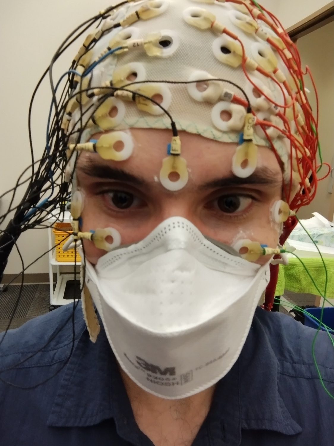 Headshot of Patrick, a white man wearing a dark blue shirt, with an EEG cap on his head. The cap has many electrodes looking like small white circular rings attached; numerous wires of various colors are connected to these electrodes.