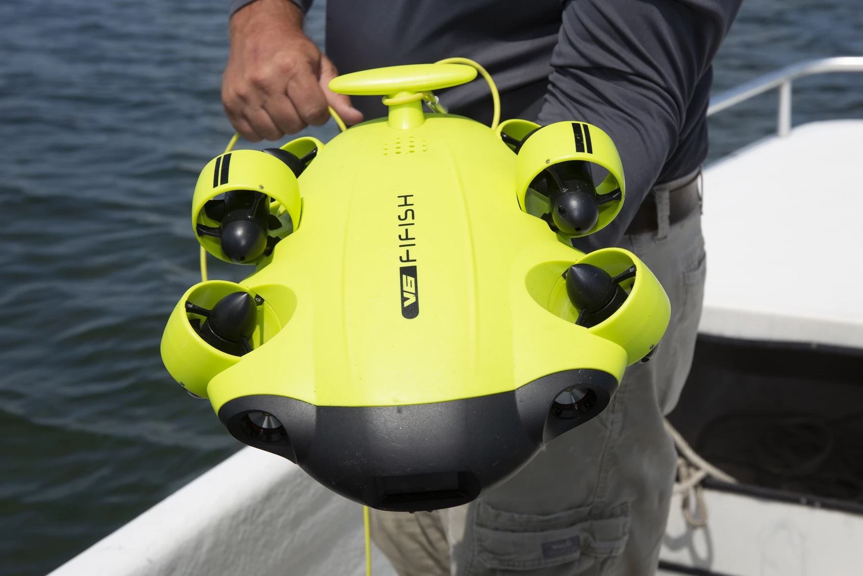 a small bulbous robot submersible, about two feet long, held by someone on a boat. it has four small propellers, two on each side, and two cameras on the front, resembling eyes on a face