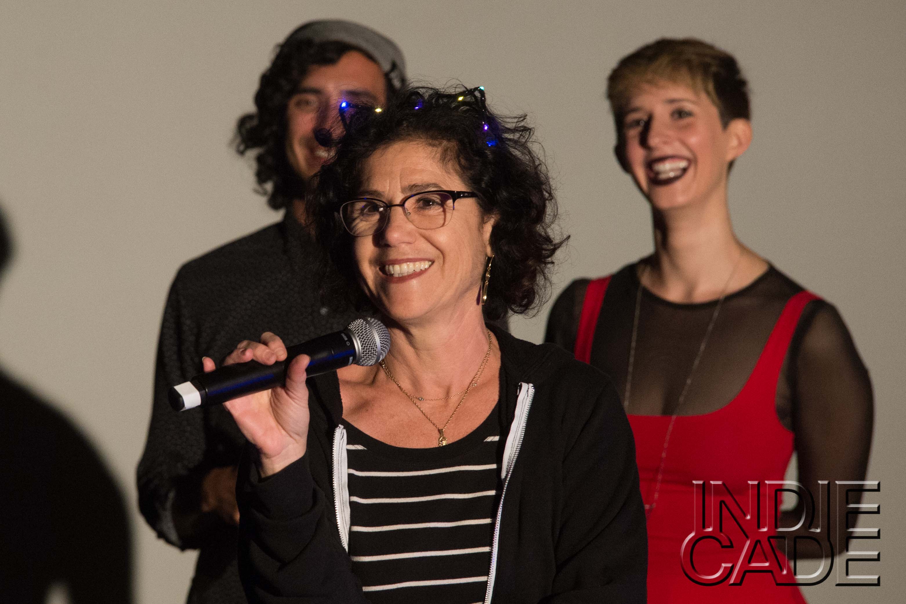 a white woman wearing glasses with sparkly led in her hair holds a microphone and smiles. Two other white people stand behind her smiling