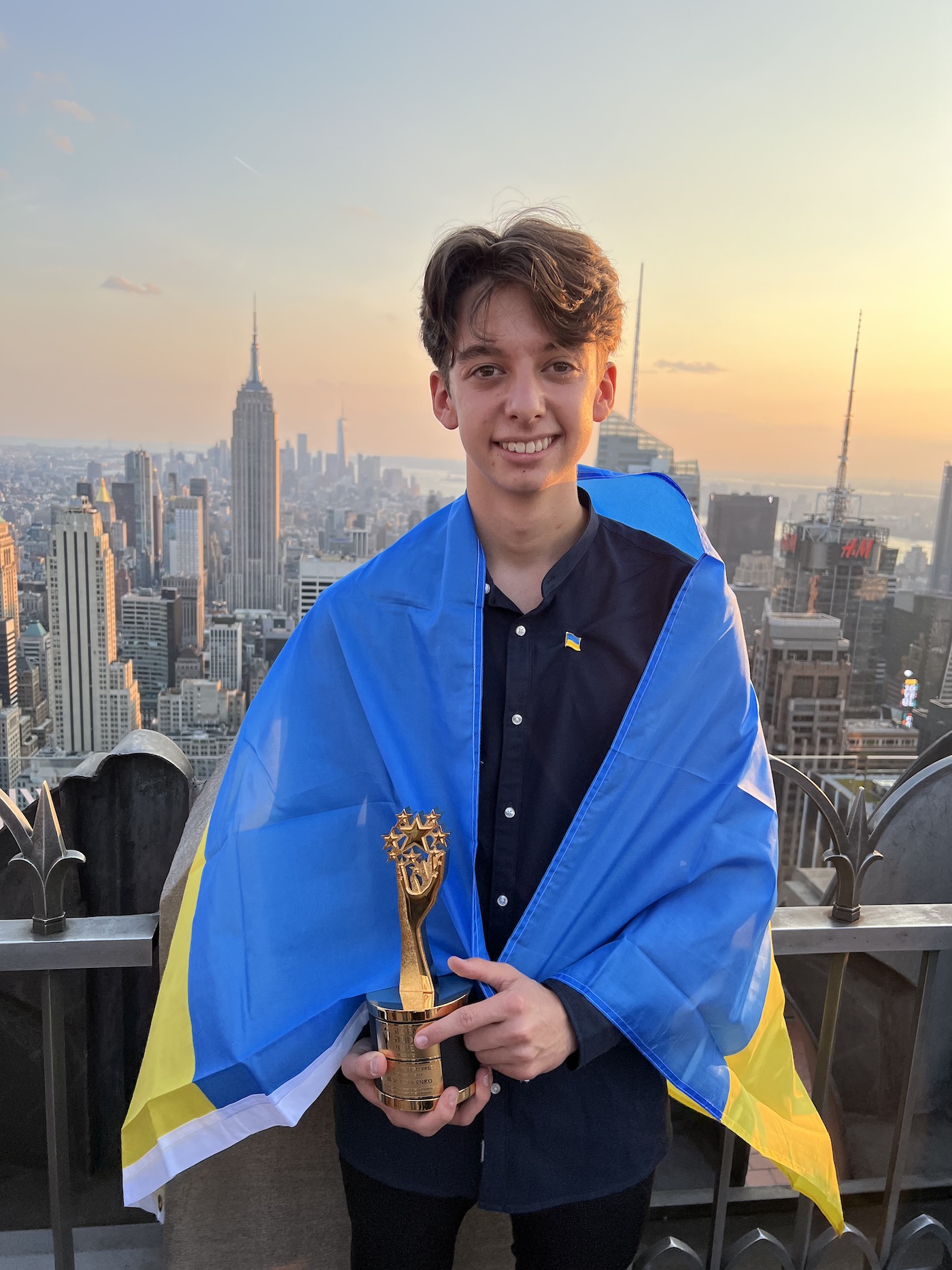 A smiling teen holding a trophy looking into the camera, wearing a Ukranian flag around his shoulders with the NYC skyscape in the background.