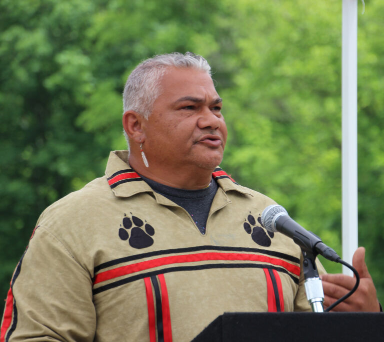 A man speaking into a microphone. His beige shirt has two animal pawprints on the left and right of his collar.