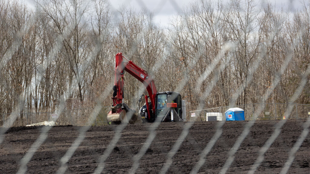 A red crane pulls at dark soil from behind a chicken wire fence. Barren trees in the gloomy winter background.