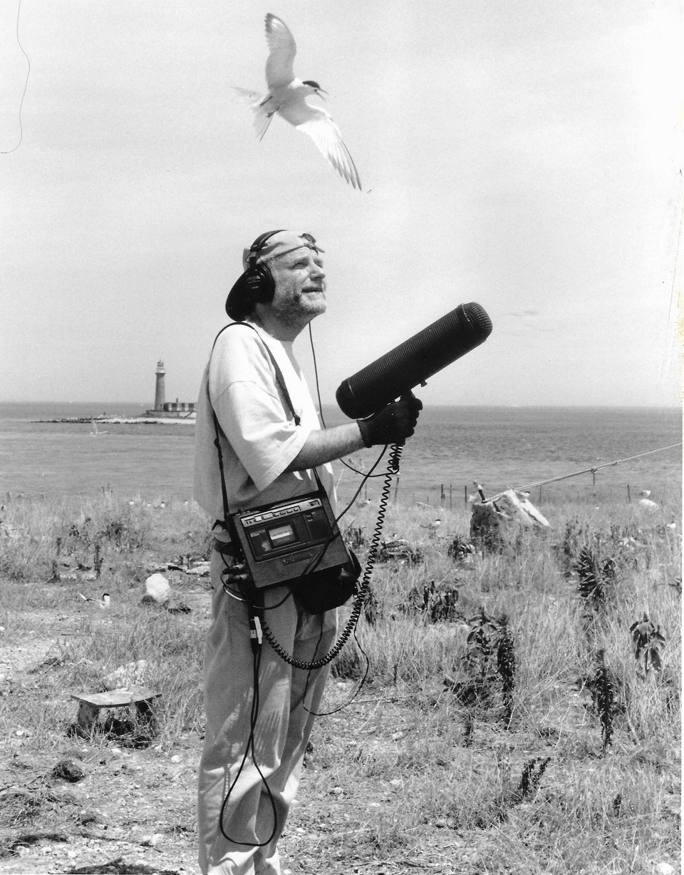 a black and white photo of an older white man wearing a backwards baseball cap with headphones and a large mic recorder over his shoulder, holds a large microphone with a wind cover on it as a seagull flies over his head