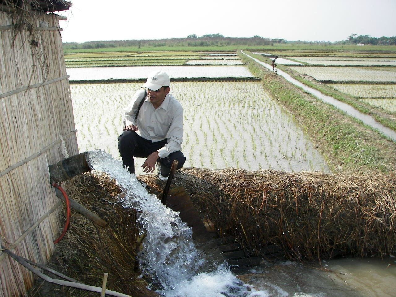 a bangladeshi farmer kneels next to a building built out reeds with a metal pipe coming out of it, which is gushing water into a man-made waterway beneath it