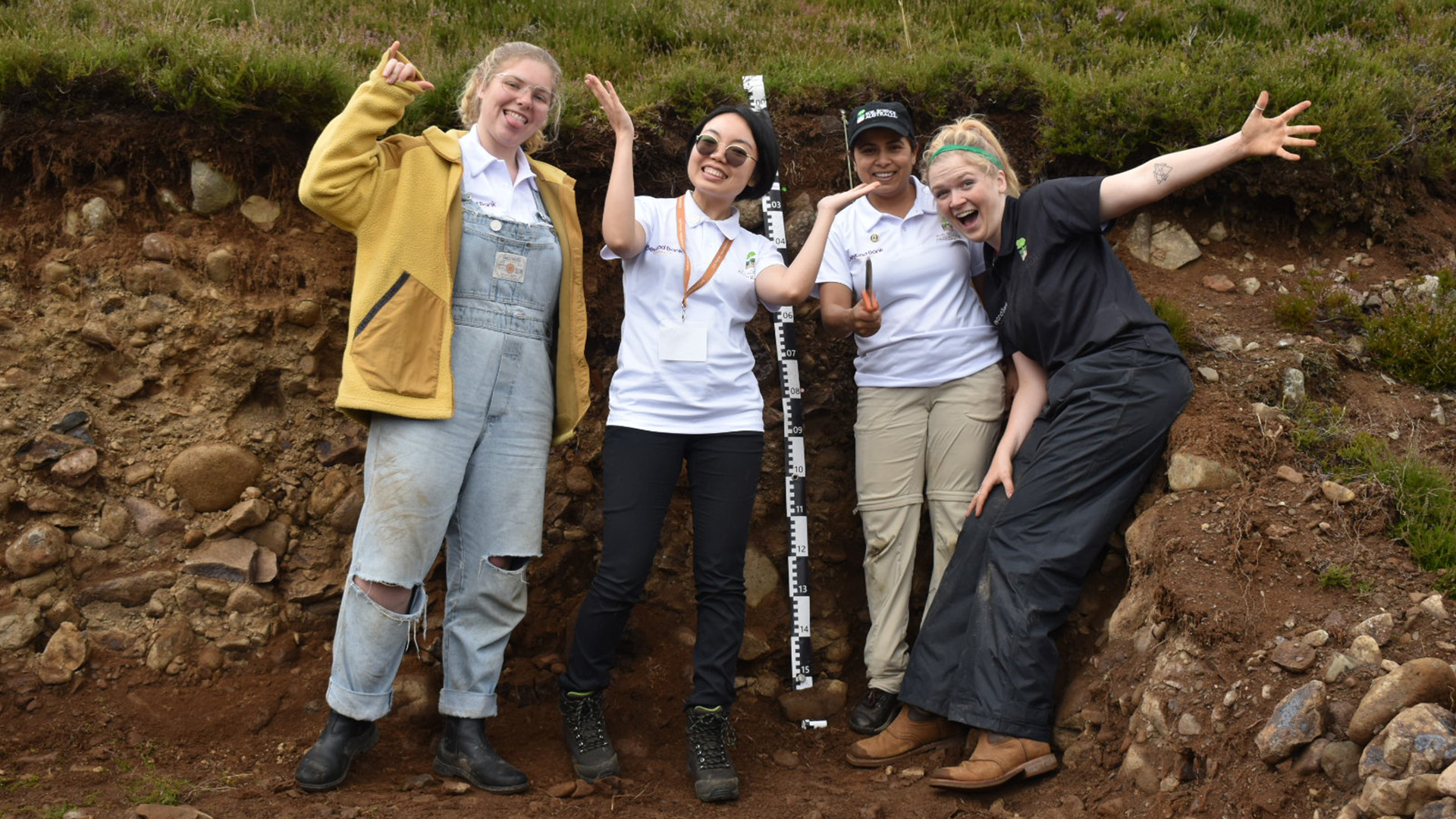 four young soil scientists stand in a soil pit with grass above and rocks around them. they are smiling with their arms up in the air in various ways, and look extremely joyful