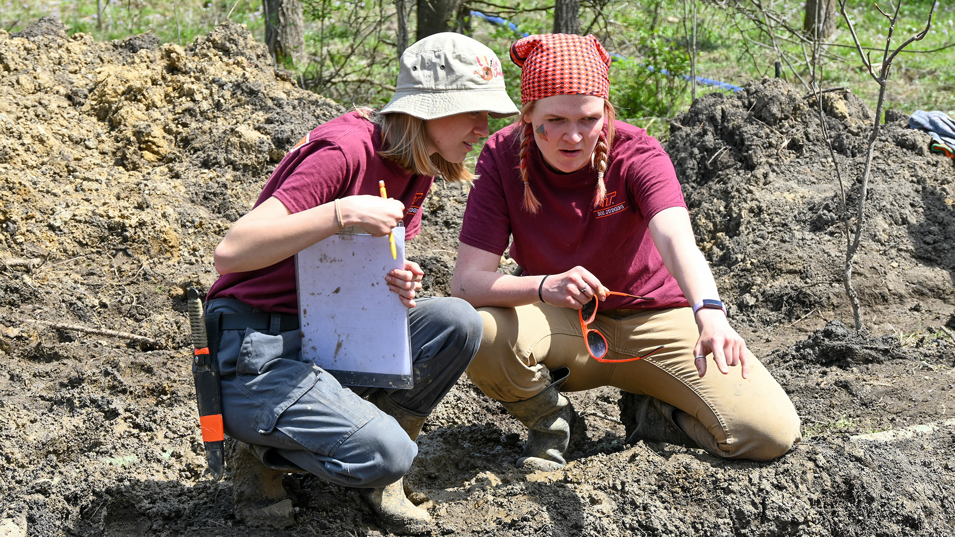 two women kneel in a muddy, dug-up area and are looking together at a soil pit, deep in concentration