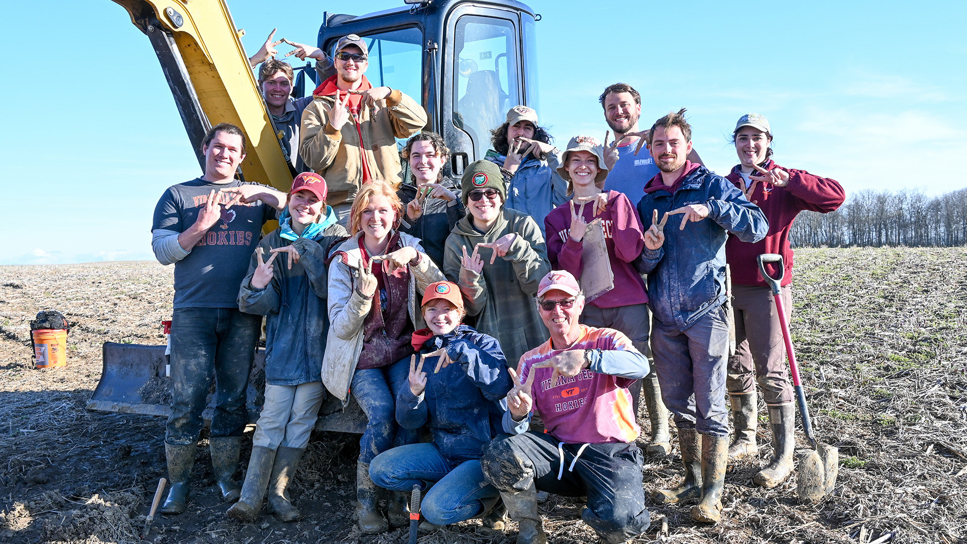 a group of fourteen people gathered in front of a backhoe machine, smiling at the camera with their clothes covered in soil