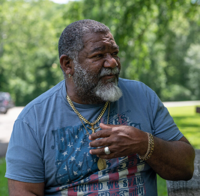 A middle-aged man with an American flag t-shirt looking to the right, his fingers touching his gold necklace.