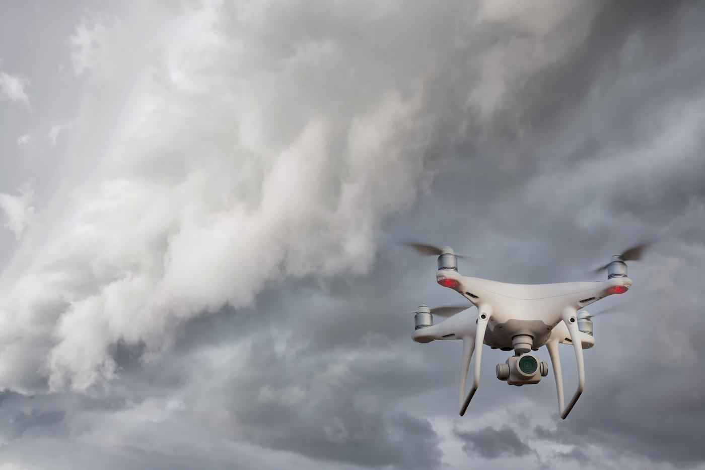 A white flying drone with a camera facing the viewer flies on the lower right hand corner. In the background, dark grey storm clouds are forming.