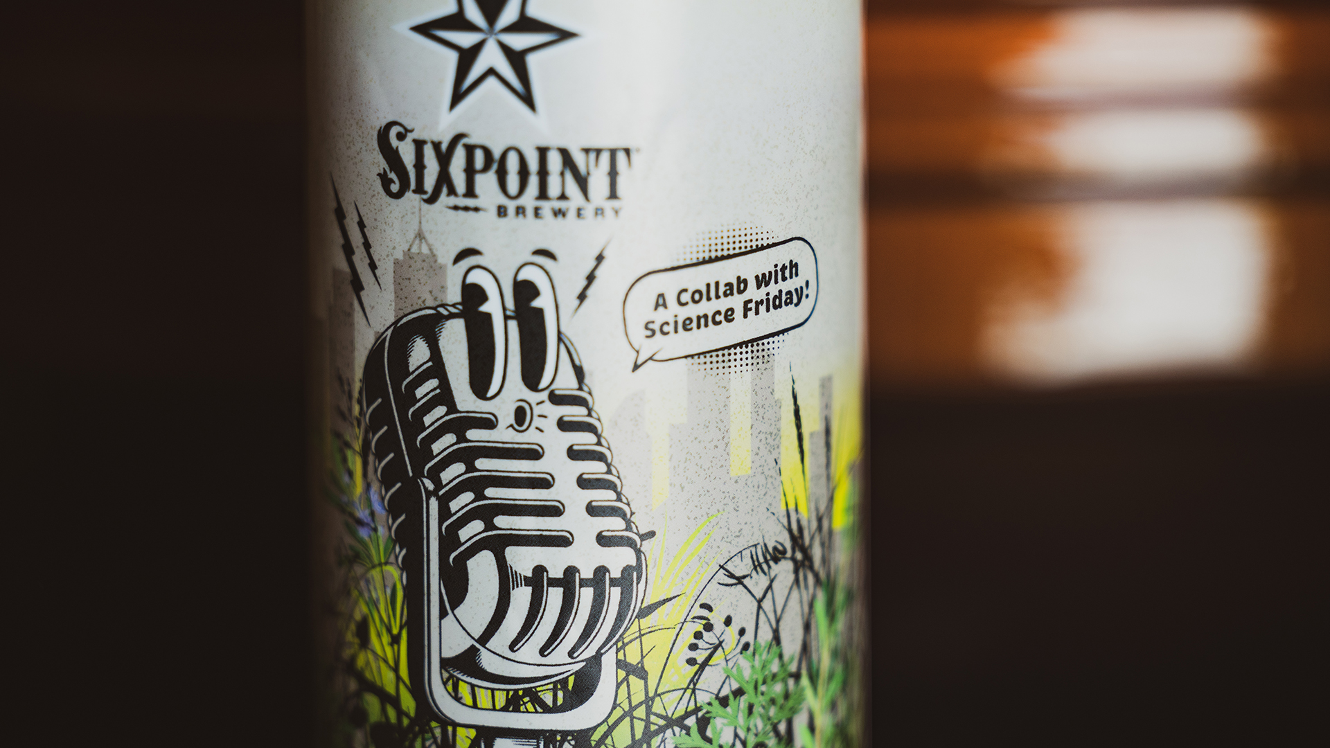 a closeup of a beer can. on the label is a old-school microphone with cartoon eyes looking to the left, it is also whistling. Behind it is a field of plants and a city skyline. the can says 'sixpoint brewery' and a speech bubble coming from the microphone says 'a collab with science friday!'