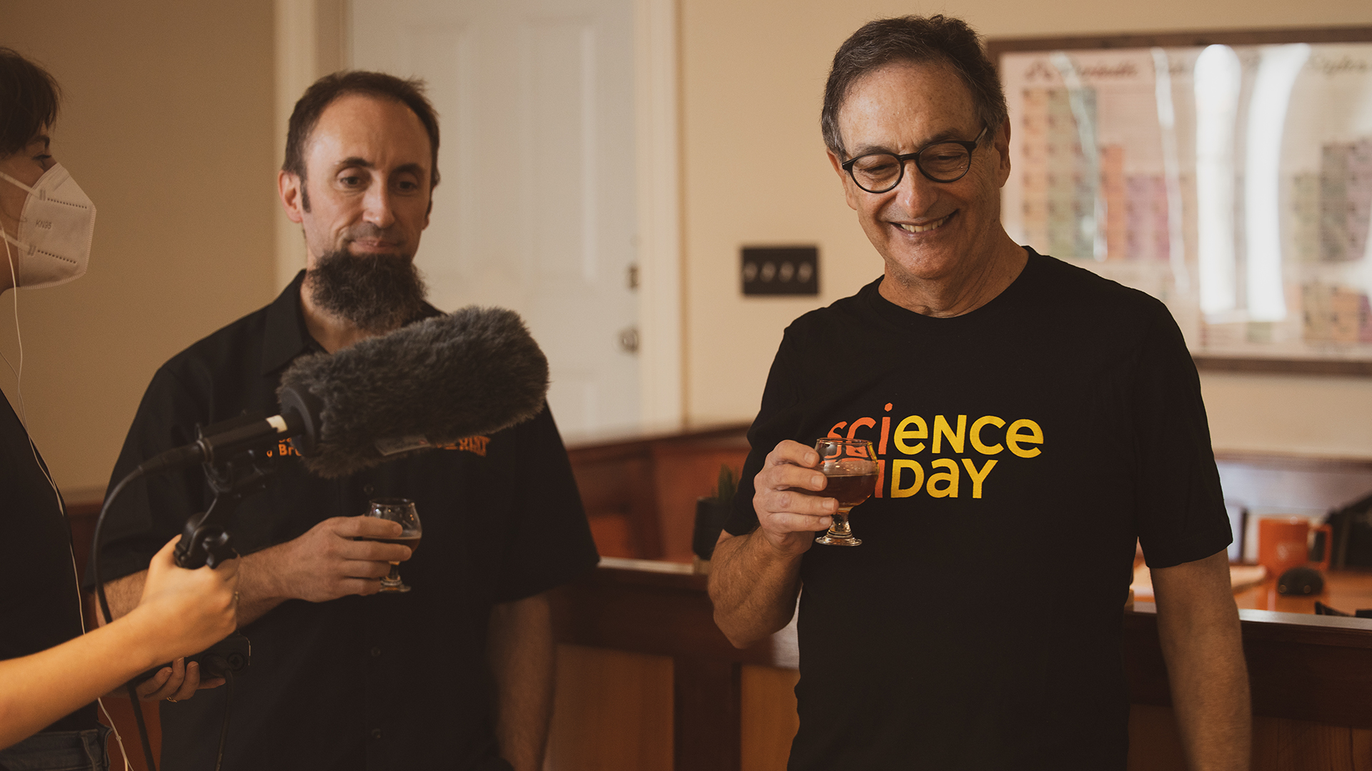 an older white man holding a small beer glass wearing a black t-shirt that says 'science friday.' next to him stands a white man with a large goatee. a woman to the left wears a mask and holds a large microphone pointed at them