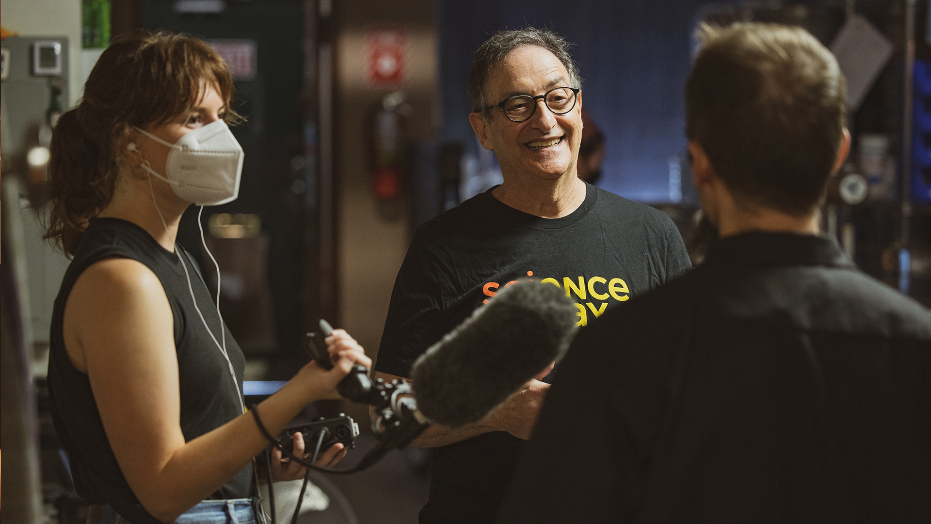 a young woman wearing a mask holds up a microphone to man, we can only see his back. standing in front of him is an older white man laughing