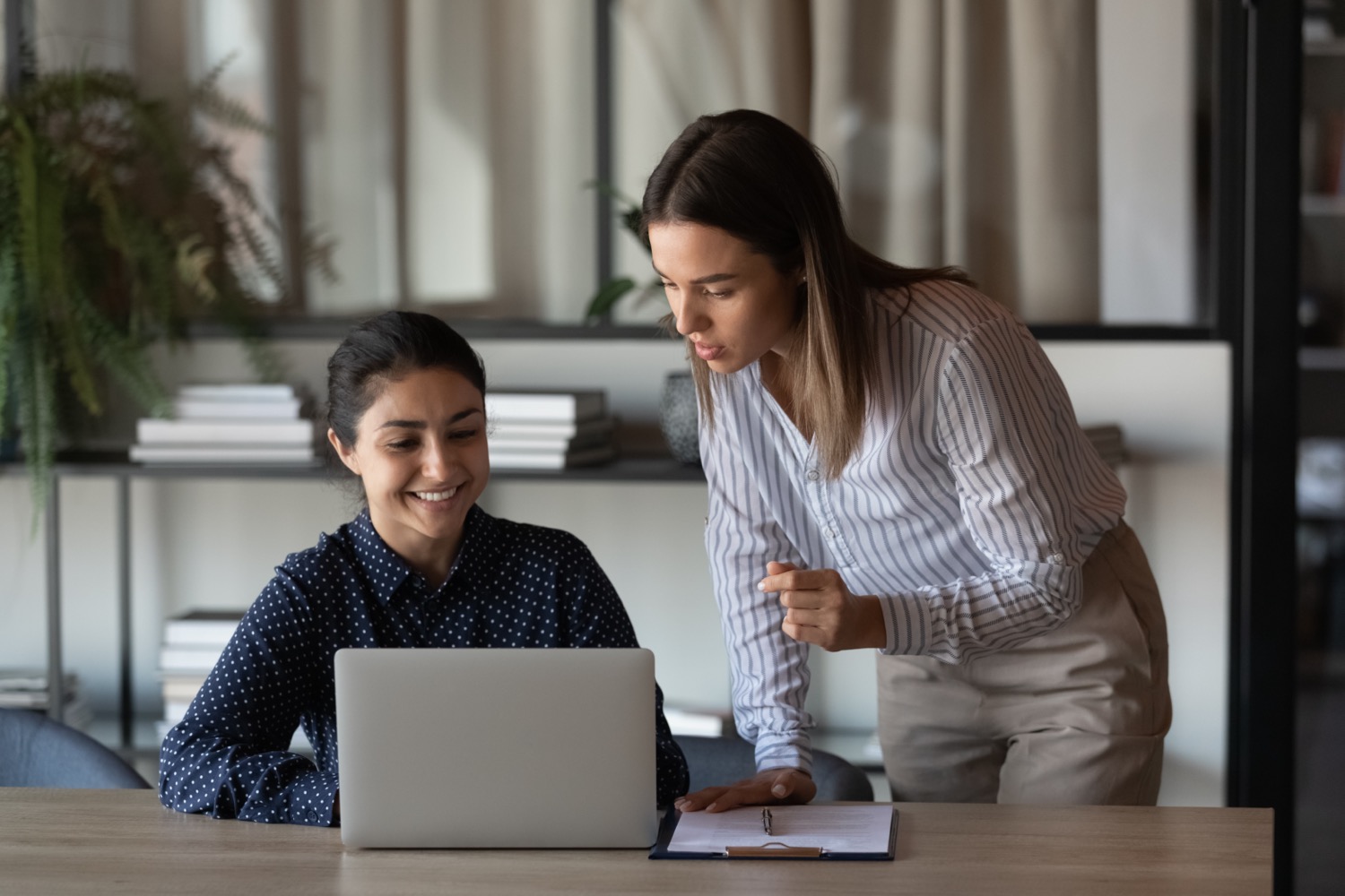 two women in an office. one on the left is sitting down and smiling at a laptop, and the other woman is standing to her left leaning down gesturing at the laptop