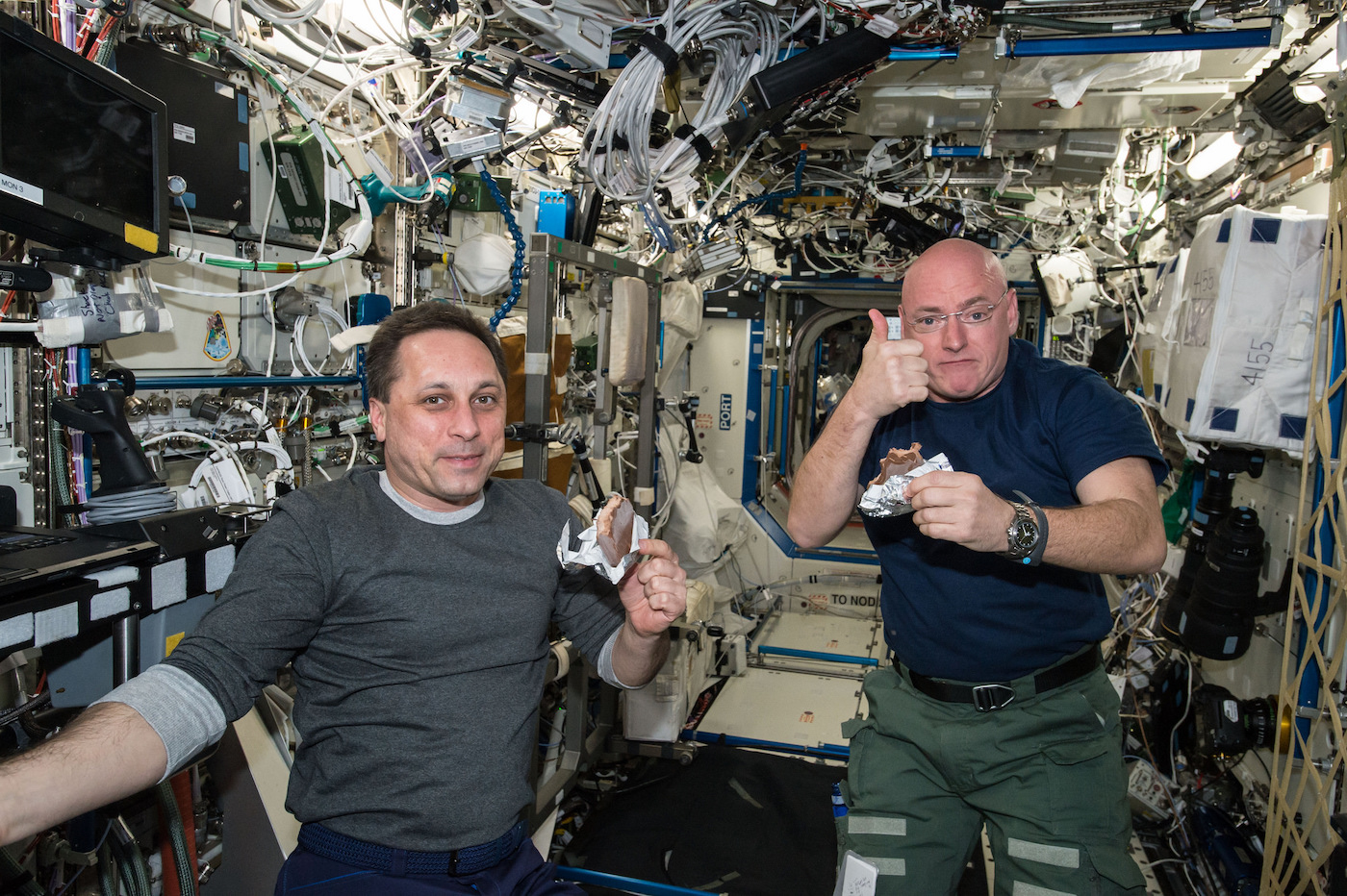 two men float in 0 gravity aboard the international space station, in a room full of pipes and wiring.