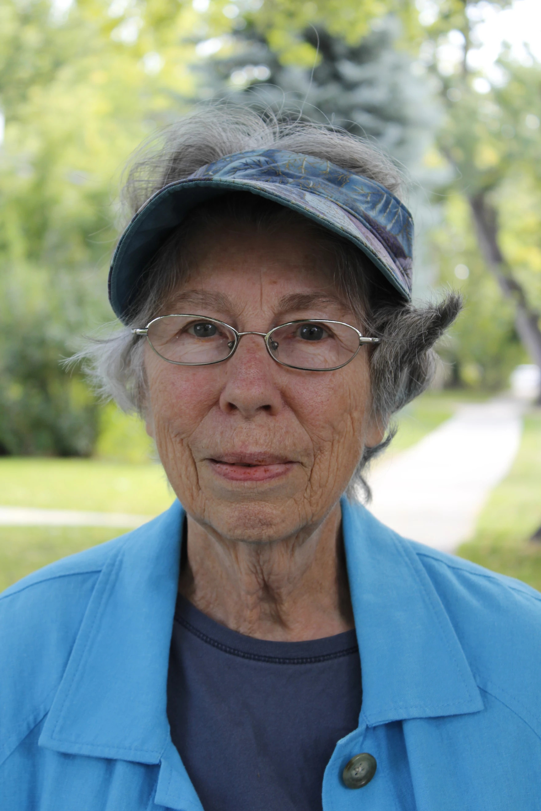 an older white woman, wearing glasses, a hat, a light blue jacket, and a darker blue shirt looks penisvely at the camera outside, with trees behind her