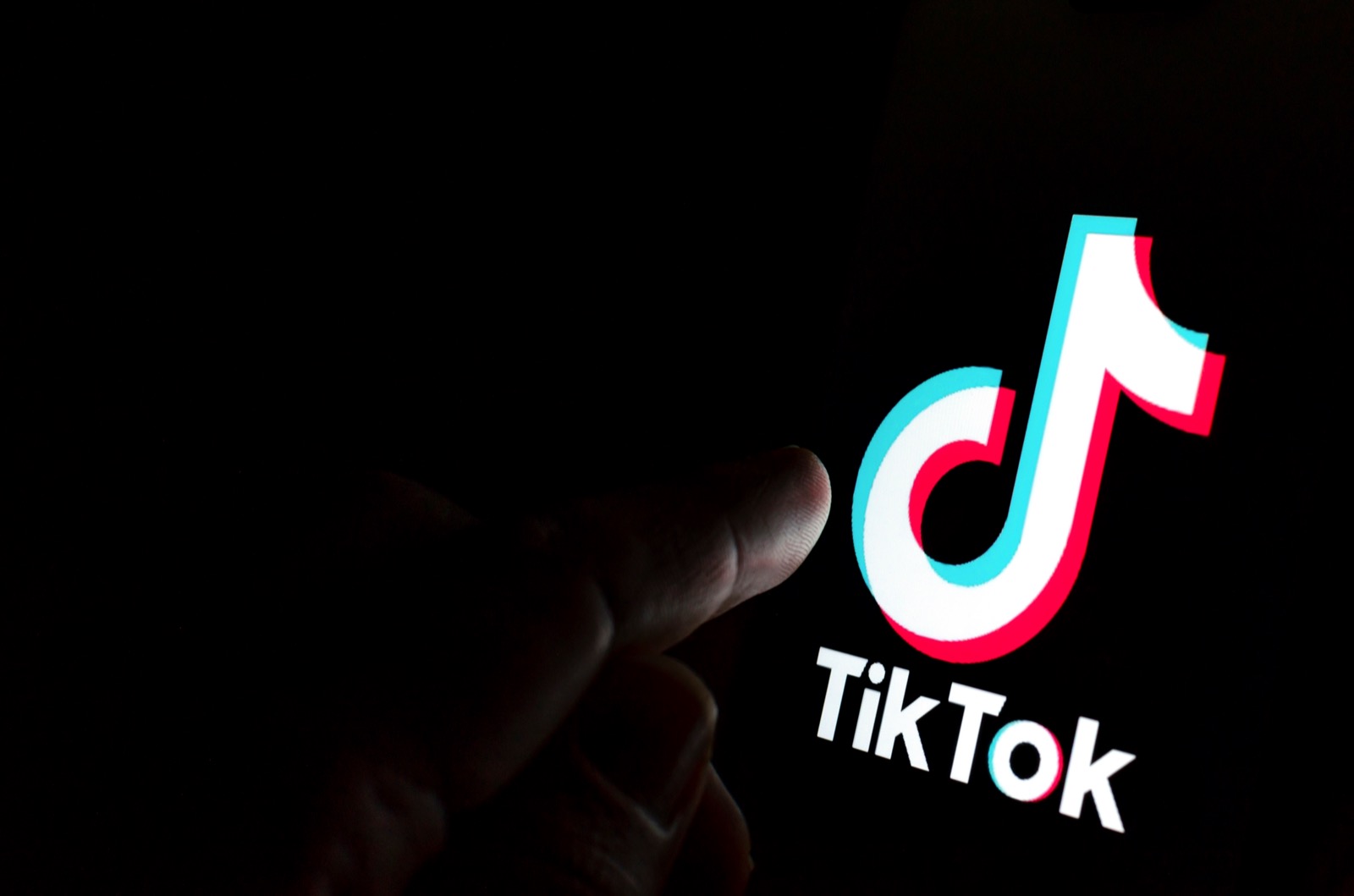 an index finger reaching out of the inky blackness to touch a tiktok logo that's only illuminating the tip of said finger