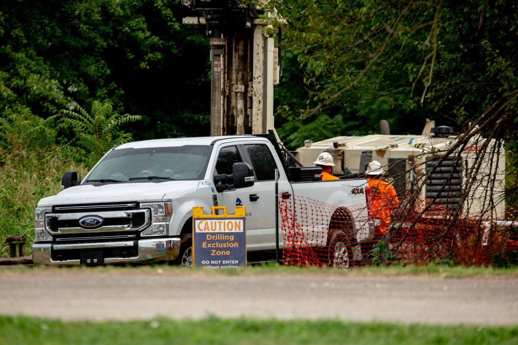 A pickup truck on the side of a road with a few construction workers surrounding it. Forest background.