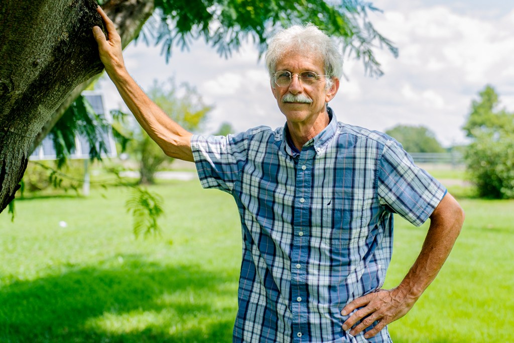 A man stands with his arm on a tree, green grass hills in the background