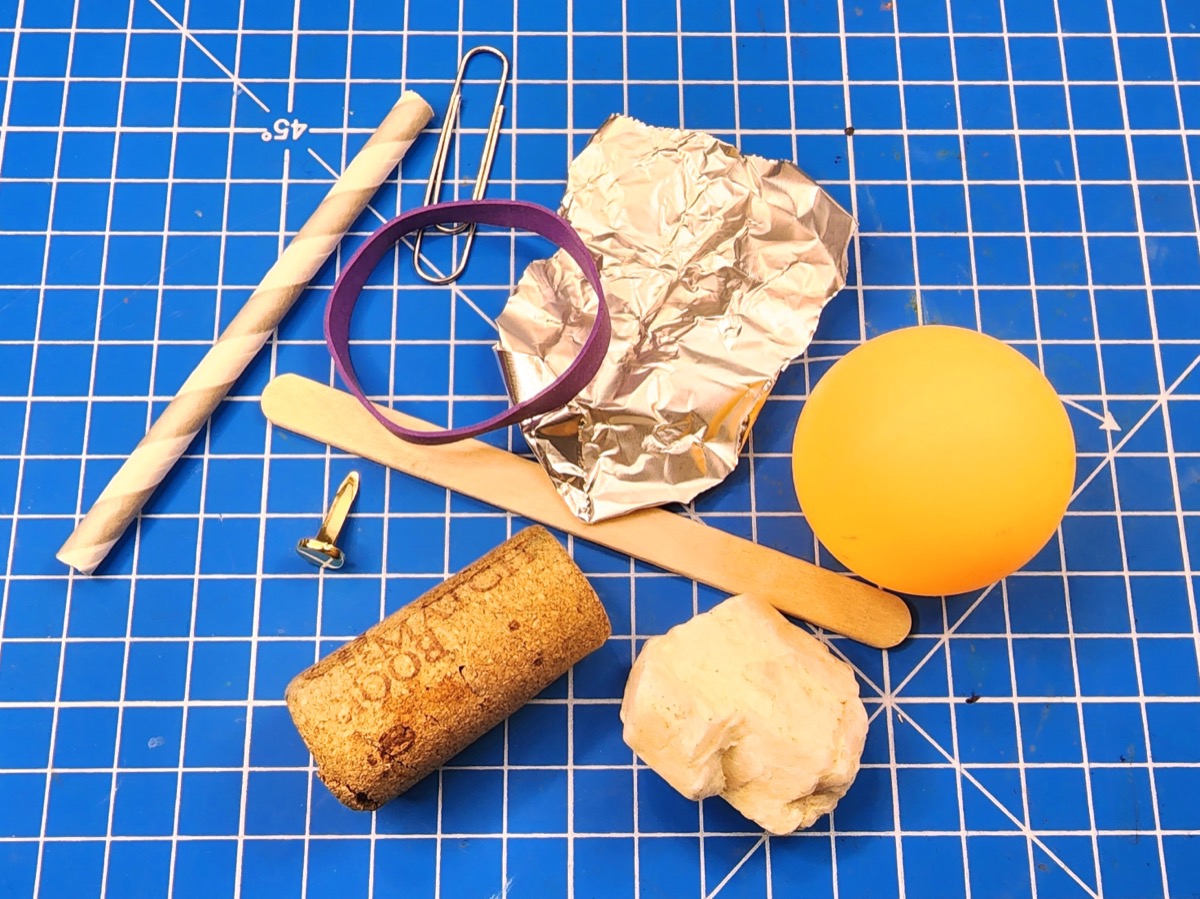 A picture of various items, including a ping pong ball, a popsicle stick, a piece of foil, a paper clip, a rubber band, a paper straw, a cork, a rock, and a brass fastener.