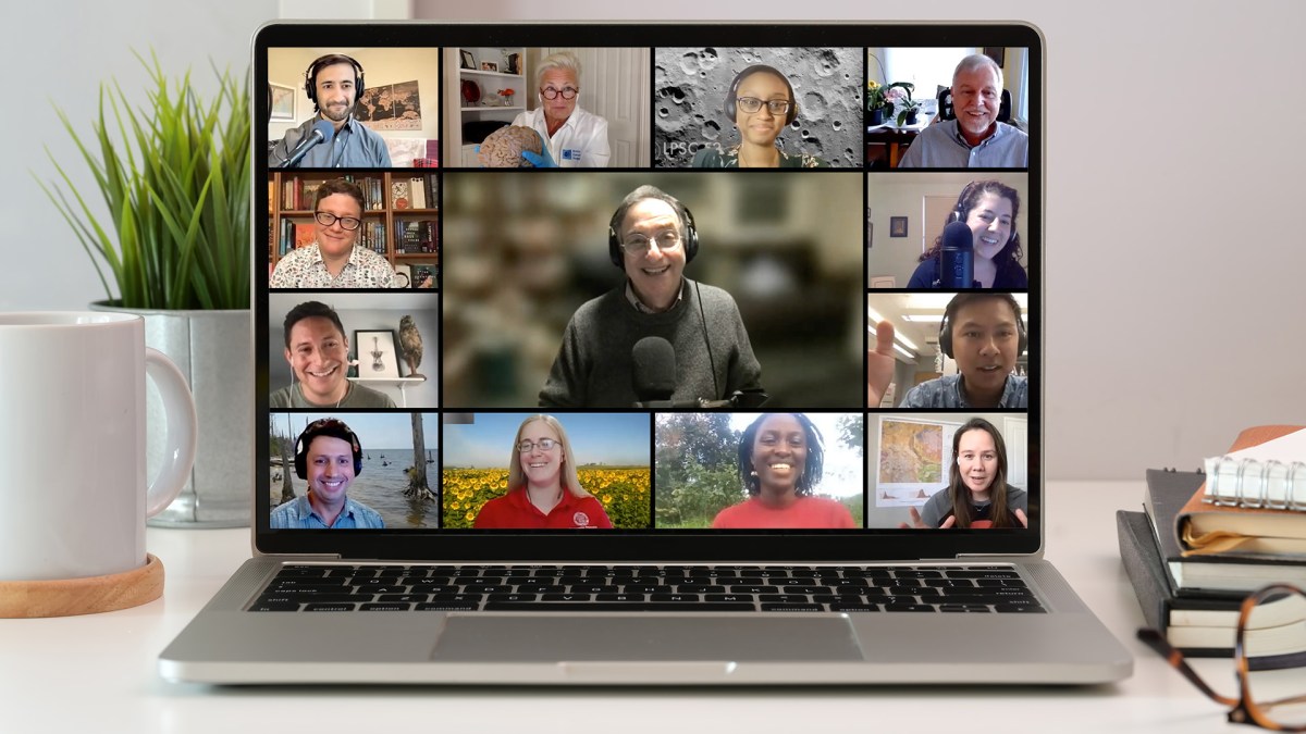 a photo of a laptop open on on desktop with a mug, plant and books. on the laptop screen is a tiled image with several smiling faces, like a livestream event.