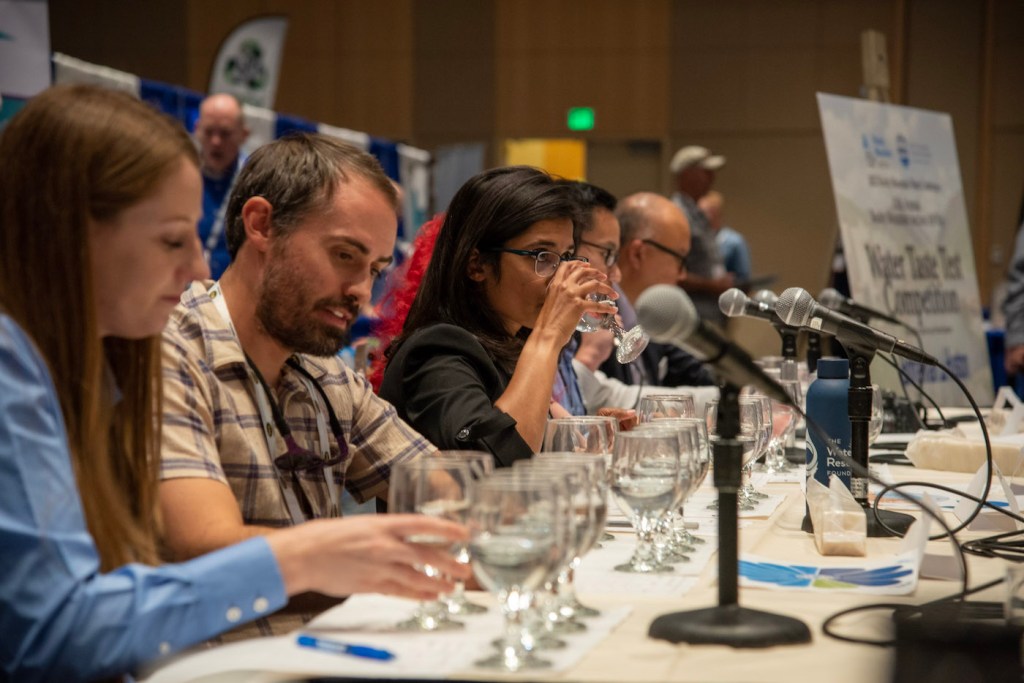 A panel of seated people, one woman in the middle is taking a sip of water.