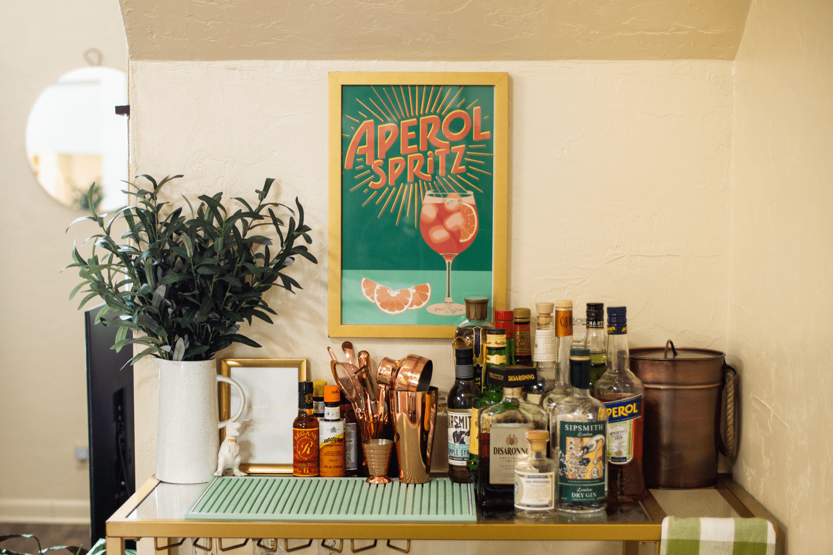 a home bar cart, with a small plant on the left, a tiny ceramic dinosaur and a bevy of cocktail mixers and utensils, alongside bitters and liquors. above the cart is a gold-framed 1920s style poster of a wine glass filled with aperol spritz, an orange slice, and some ice cubes with the words "aperol spritz" above. some orange slices sit next to the glass. 