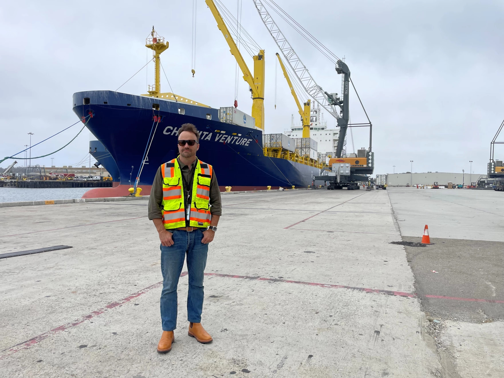 a white man wearing a hazard vest standing in front of a large container ship in port