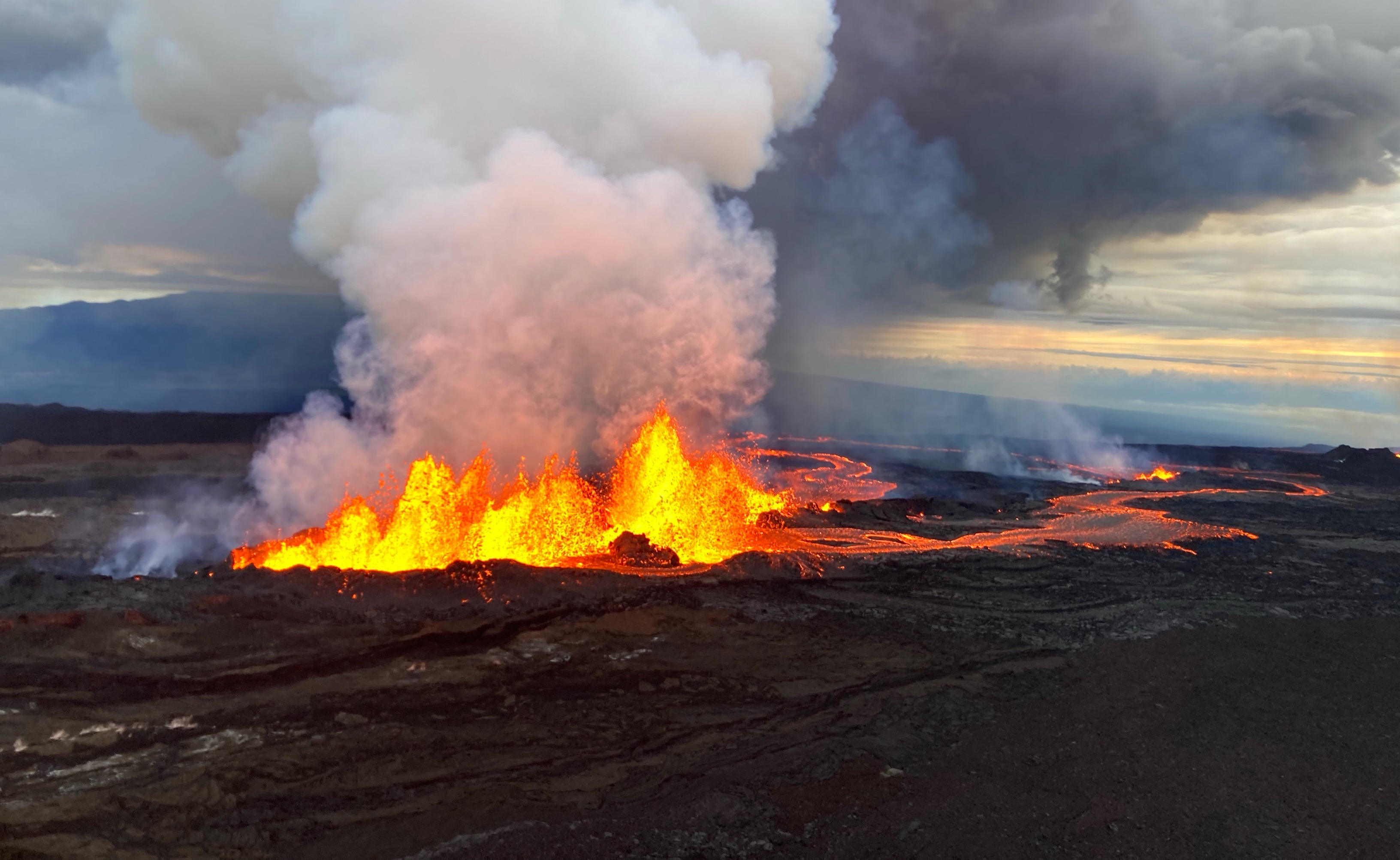 an aerial photograph of a volcanic eruption happening on a relatively straight plane of dark earth. the flames and magma from the eruption spew white smoke that turns black as it billows higher up into the sky