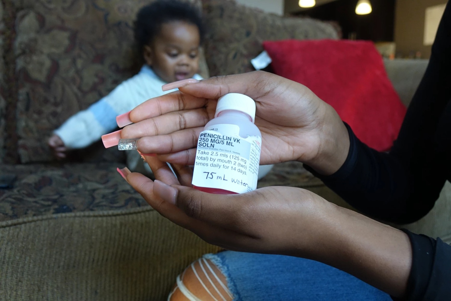 hands holding up a penicilin bottle with a baby in the background sitting on the couch