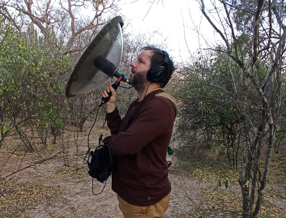 A man, Juan Pablo Culasso, holds a large metal dish towards the tops of trees, listening carefully to bird sounds.