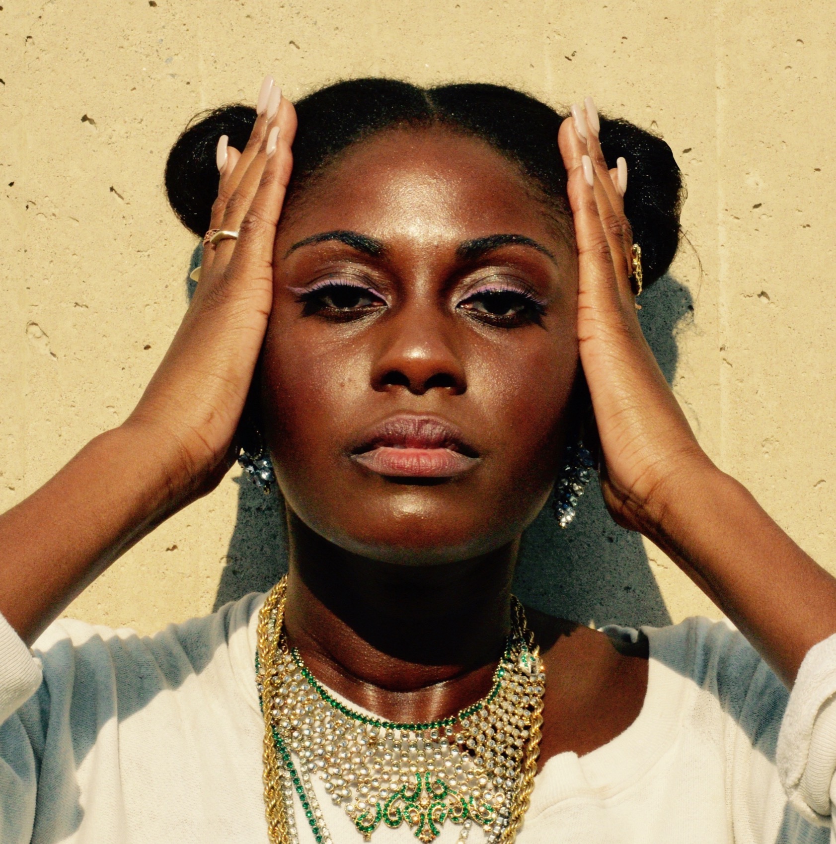 a Black woman against a wall holding her hands to the sides of her face looking directly at the camera