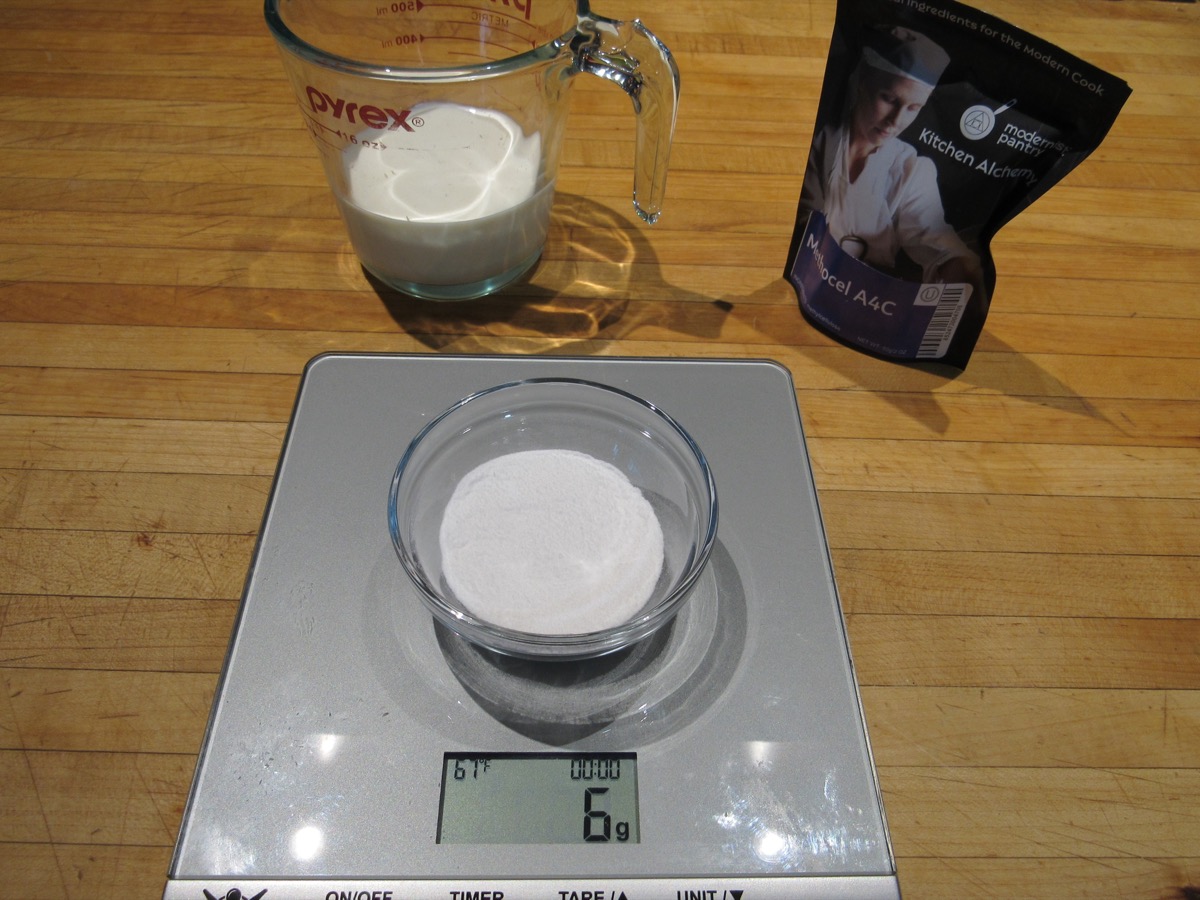 A gray digital scale with a glass dish hat holds a white powder. Behind them is a glass measuring cup filled halfway with cream, and a black bag of Methocel A4C. 