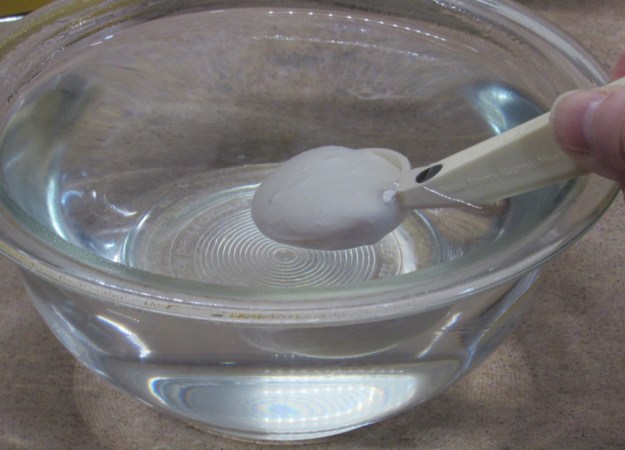 A white plastic measuring spoon holds the methyl cellulose powder. It is lowered into a glass bowl of hot water.