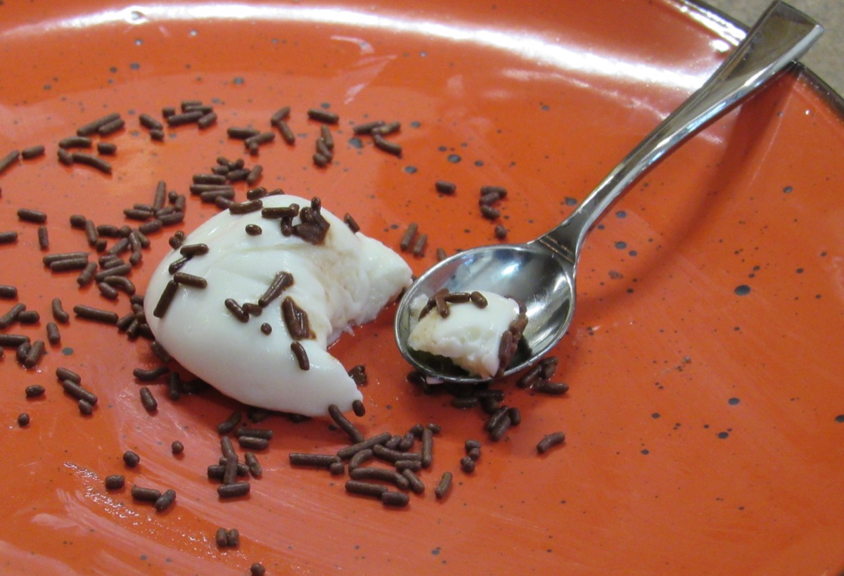 Roughly a tablespoon of a white “hot ice cream” covered with chocolate sprinkles in the center of an orange plate with a spoon.