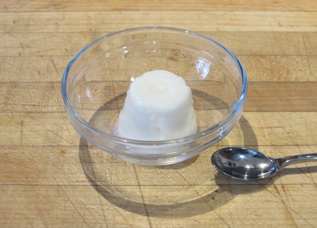 A cylindrical shape of opaque white gel sits in a glass bowl with a spoon resting to the side.