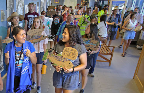 A group of people chanting and holding cardboard signs shaped like salmon that say "Our water is your skies, let us fly!"